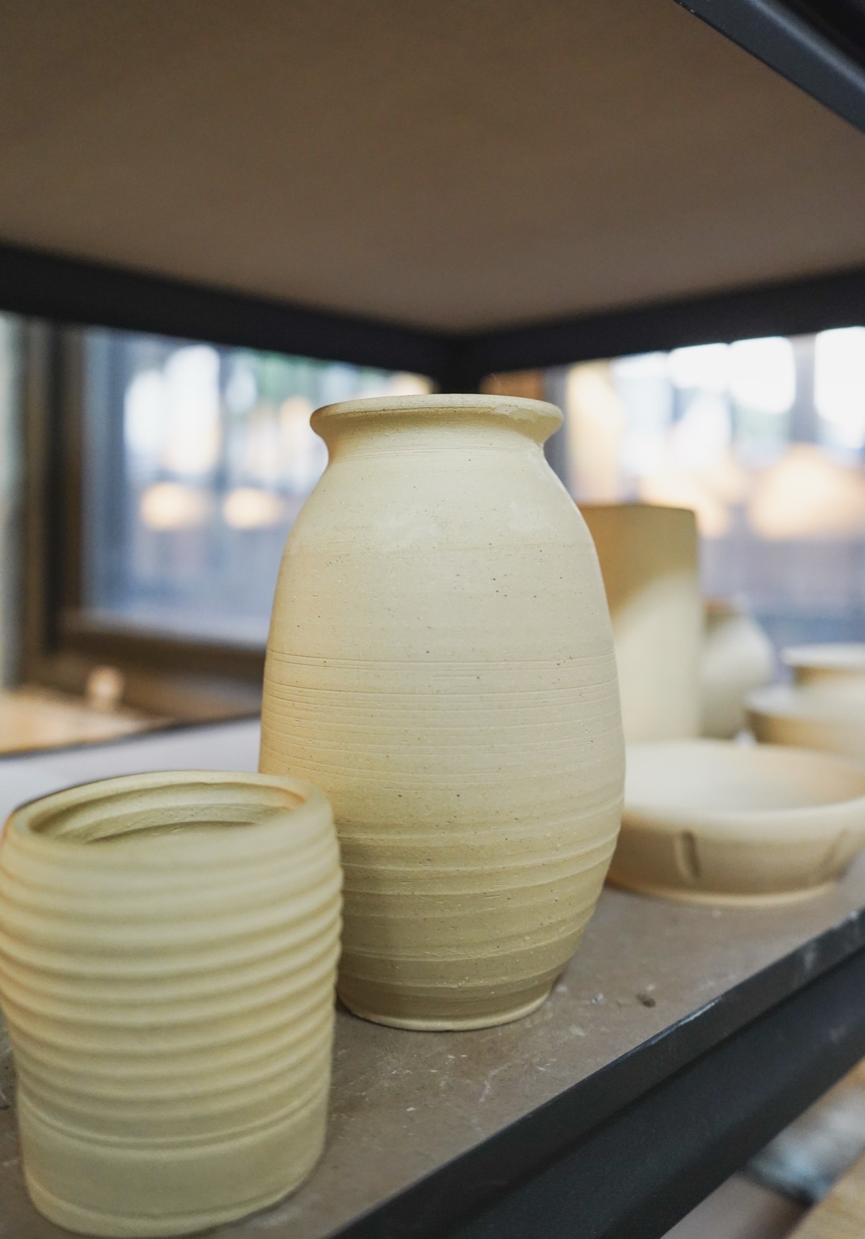 Unglazed thrown pieces drying on the shelf in window light