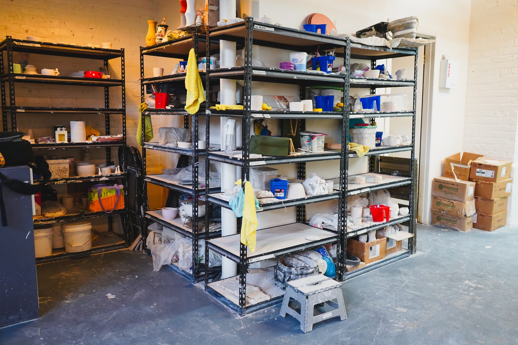 Shelves of glaze buckets and studio supplies along the whitewashed brick wall