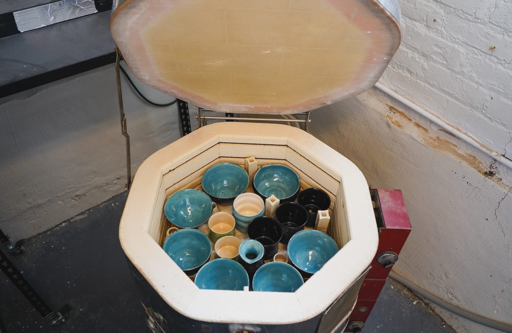 The kiln lid lifted open, revealing a full load of glazed turquoise, black, and cream bowls and cups